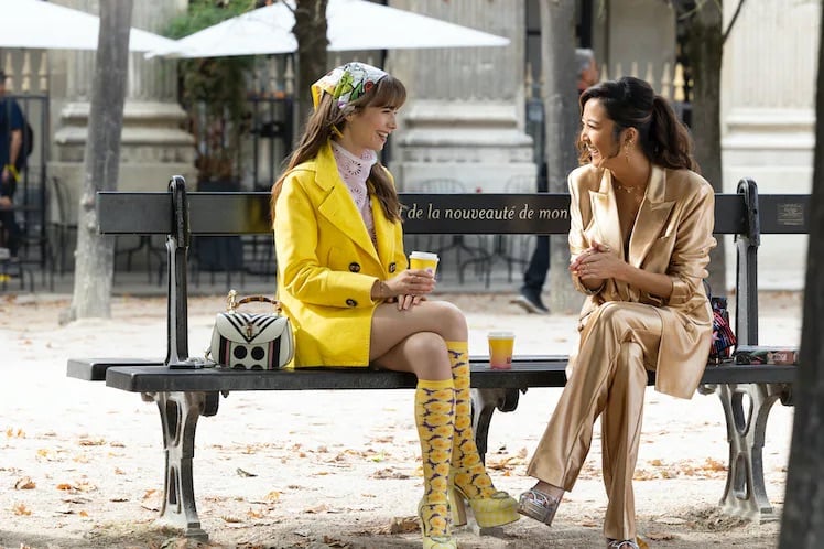 Emily in Paris Season 3 Episode 9, Lily Collins and Ashely Park are outside chatting on a bench in Paris. Lily is wearing a yellow overcoat with graphic Paul and Joe socks. Her bag is a Gucci 1947 bamboo top handle. Ashley is wearing a gold metallic suit from Rotate. | Shop the fashion and bags of Emily in Paris Season 3