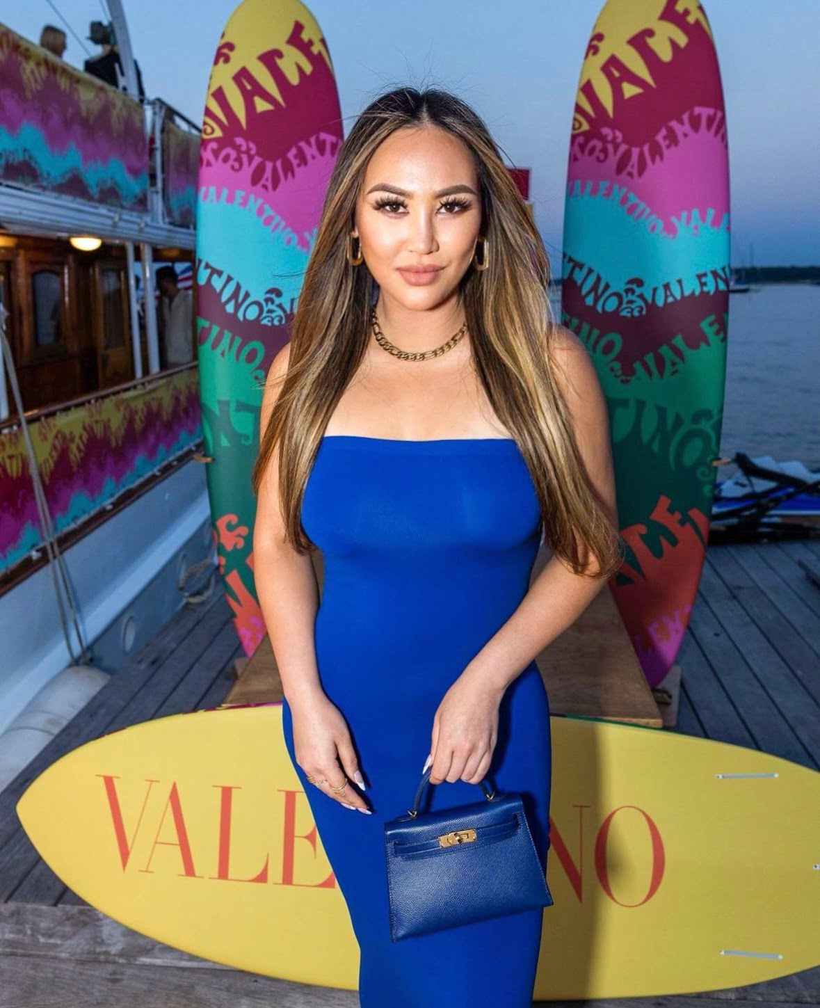 NetFlix Bling Empire NYC star Dorothy Wang at a Valentino event in front of surfboards and a boat. She is wearing a blue tube top body con dress. She is carrying an Hermes navy blue mini kelly with gold hardware.