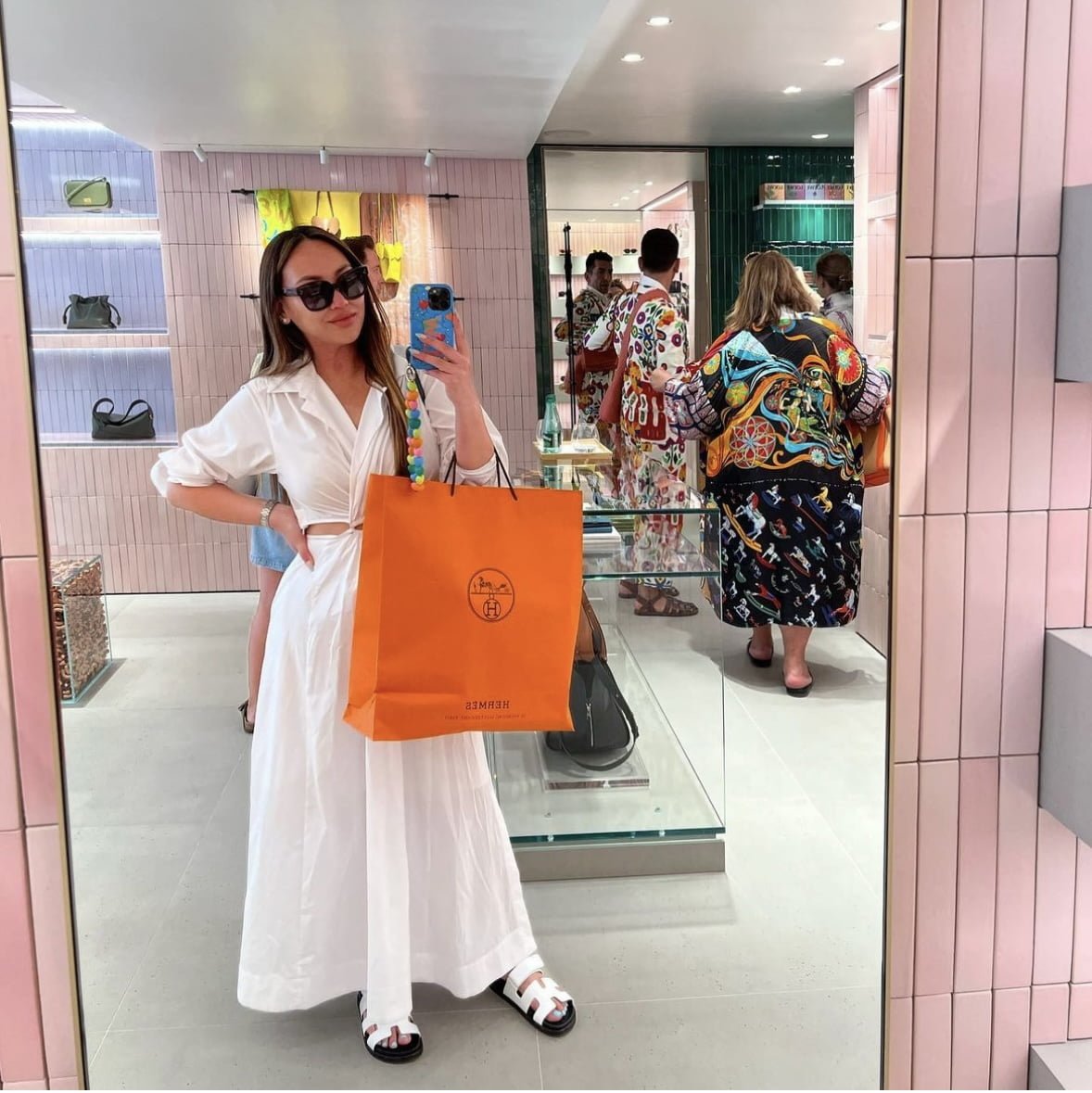 NetFlix Bling Empire NYC star Dorothy Wang is out shopping in Europe taking a mirror selfie. She is carrying a large orange Hermes shopping bag. She is wearing a white linen two piece outfit with matching white Hermes leather Chypre sandals.