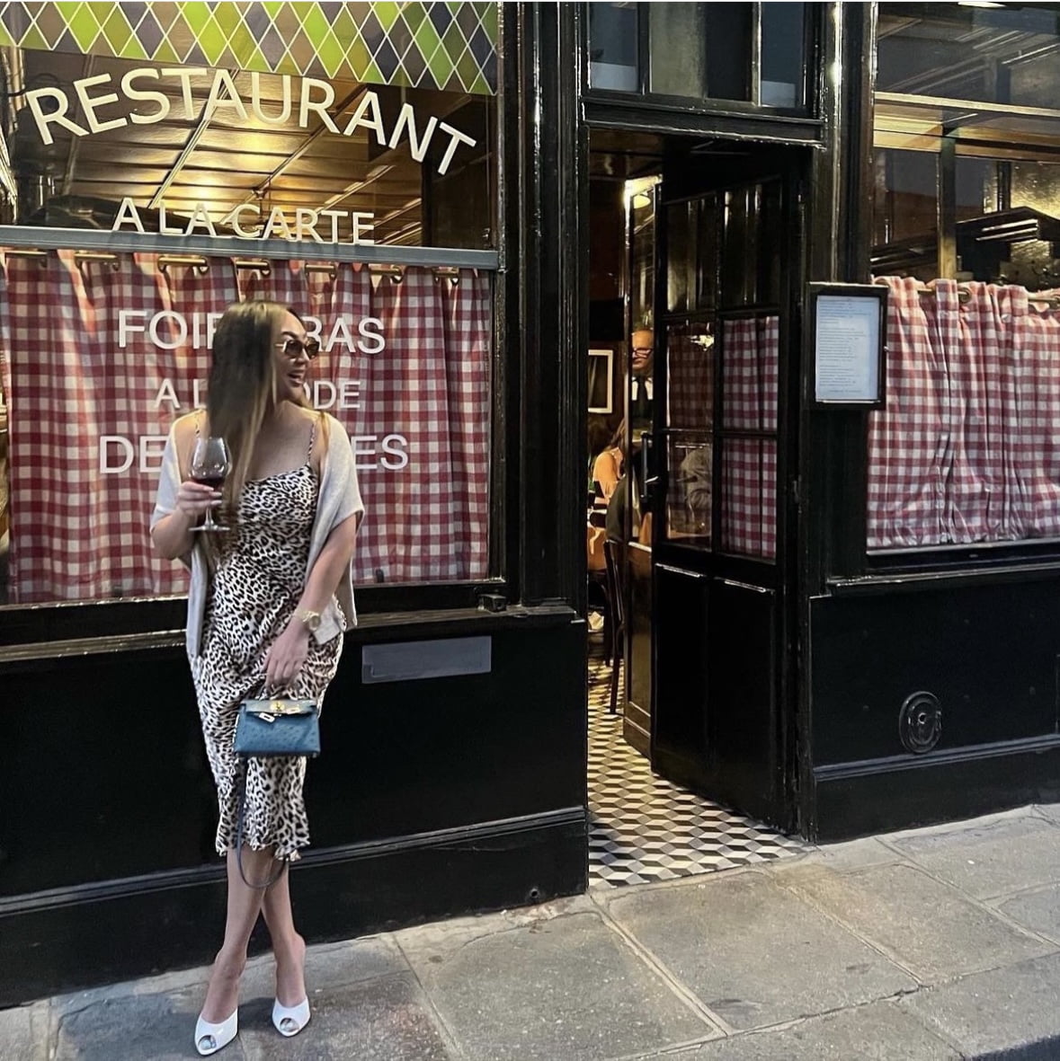 NetFlix Bling Empire NYC star Dorothy Wang wears a leopard print slip dress, white pumps and a beige sweater in front of a restaurant in Paris. She is carrying a glass of red wine and her Hermes blue ostrich mini Kelly Bag with gold hardware.