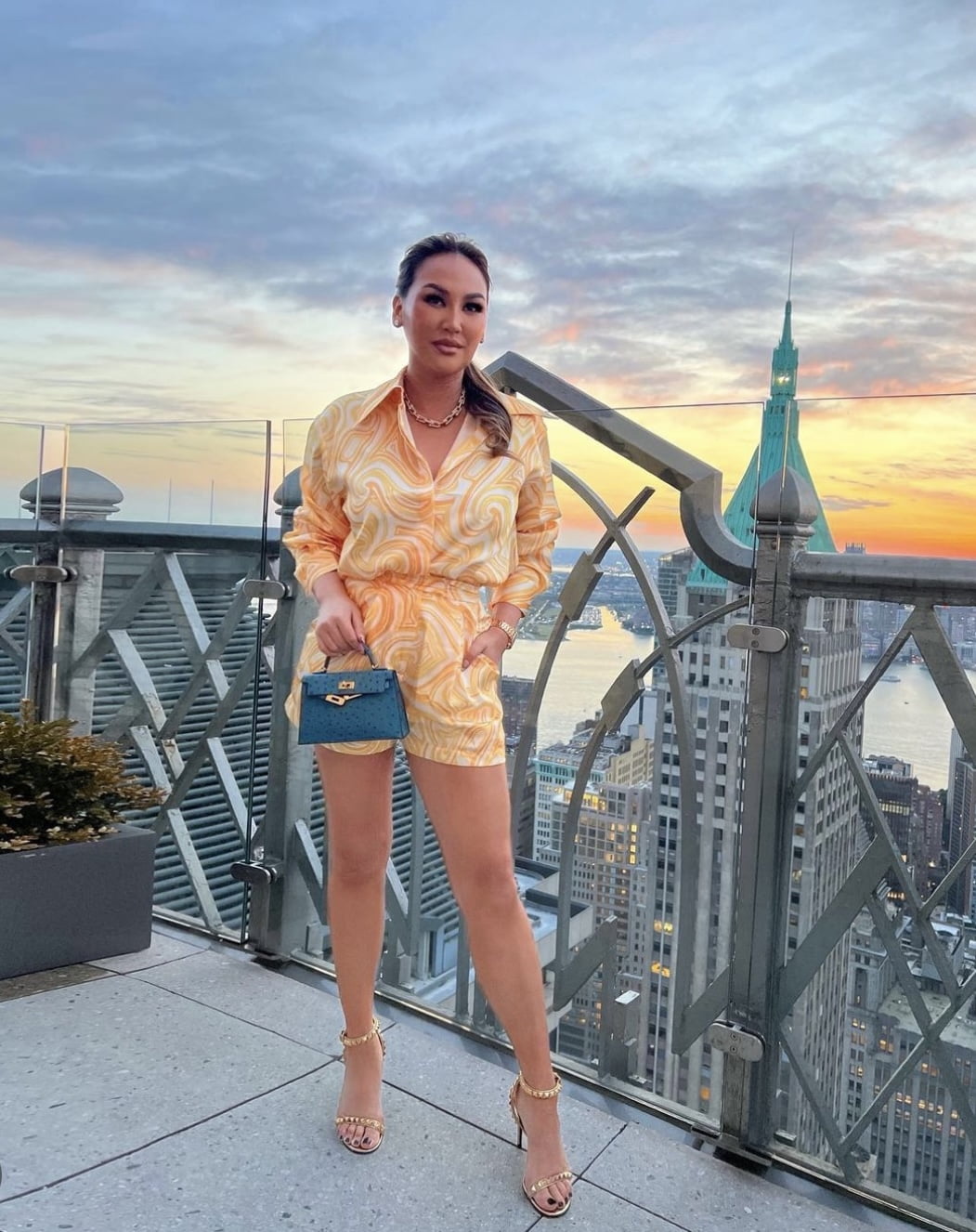 Dorothy Wang Bling Empire NYC wearing a short yellow jumpsuit. She is carrying a blue ostrich Hermes mini kelly bag on a rooftop in NYC.