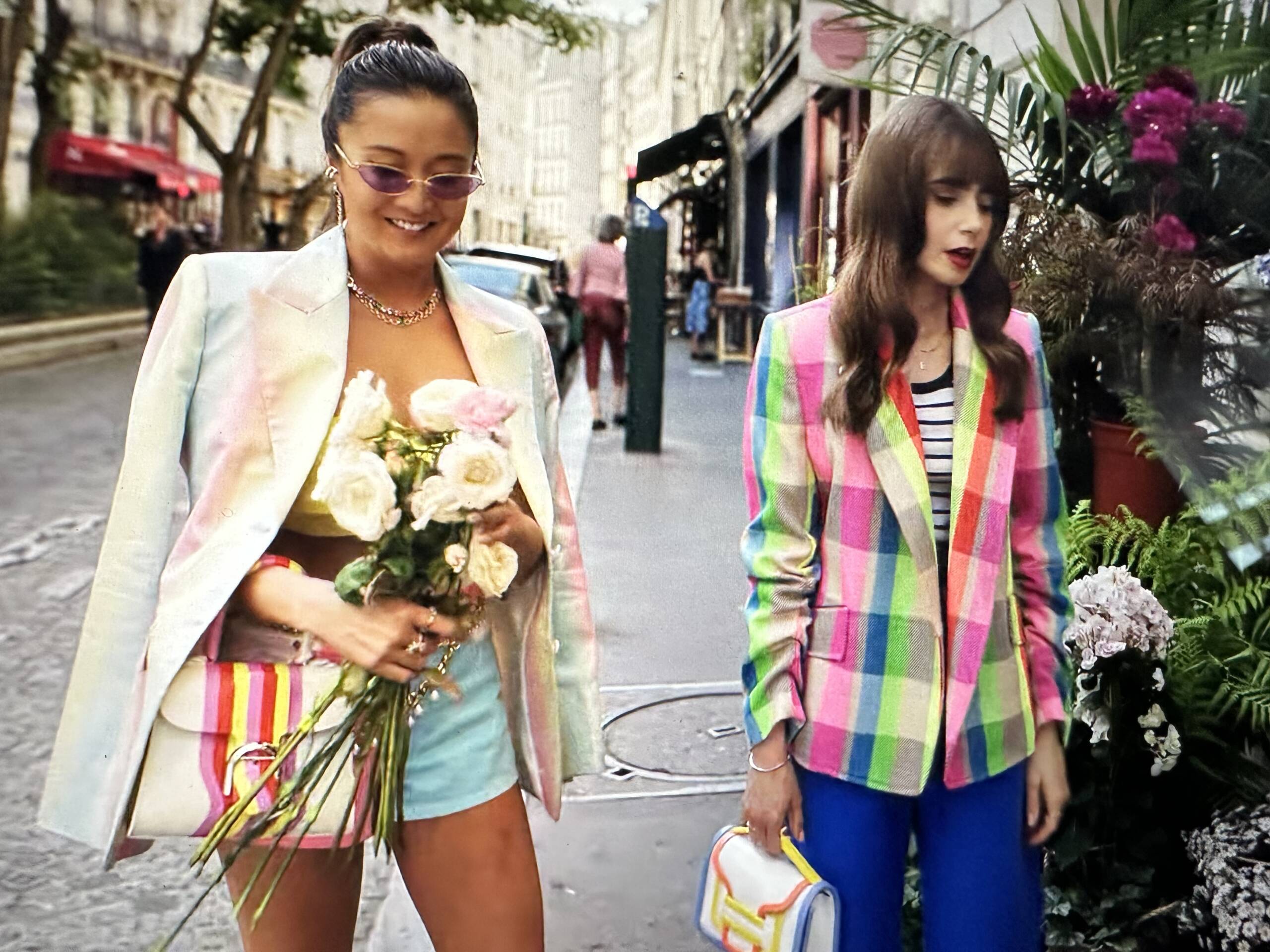 Emily in Paris Season 3 handbags features Mindy and Emily at a flower shop. Lily Collins is wearing a Mira Mikati neon checkered rainbow blazer with a Pierre Hardy Alpha Bag. Ashley Park wears a blazer with blue shorts.