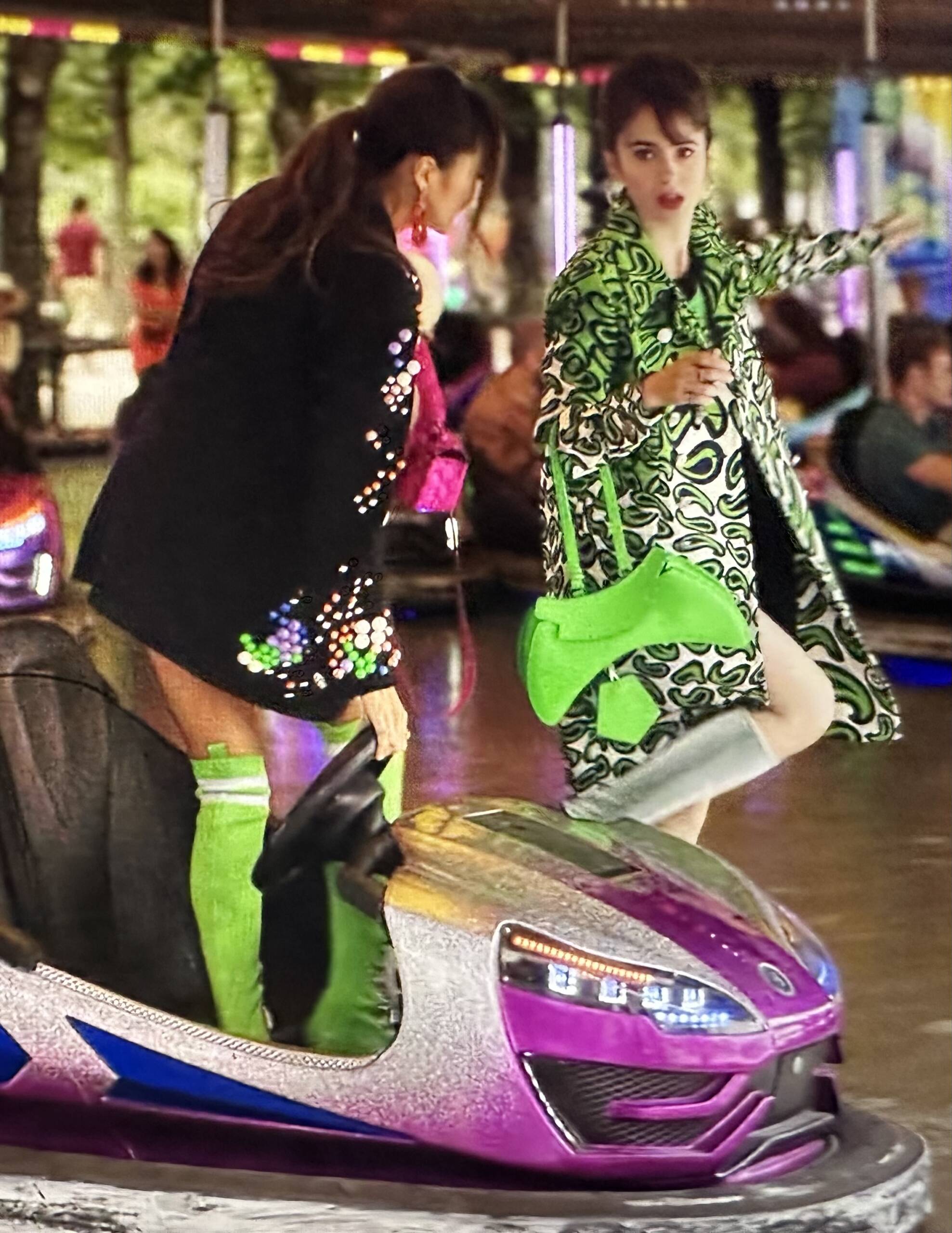 Emily in Paris Season 3 handbags feature Mindy and Emily at a the Paris amusement park in the Tullieries. Lily Collins wears a bright neon Visore X Manette bag Fluo handbag and Miu Miu printed coat. Ashley Park wears neon green boots and a sequin hot pink baguette bag | Shop Bags of Emily in Paris Season 3