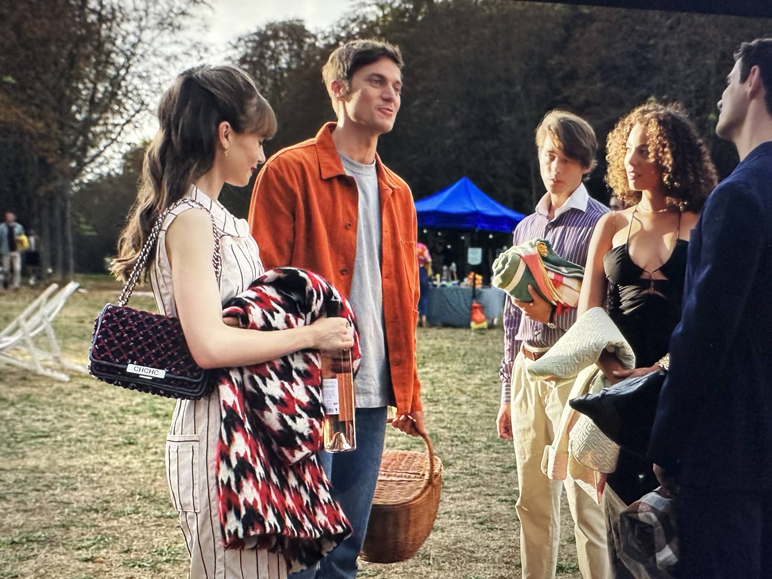 Emily in Paris Season 3 Episode 7, Lily Collins is white and black pin striped sleeveless jumpsuit with a tweed Carolina Herrera chain strap bag at an outdoor picnic with Gabriel. Her hair is in a half up-do with Birkin Bangs. | Shop the fashion and bags of Emily in Paris Season 3