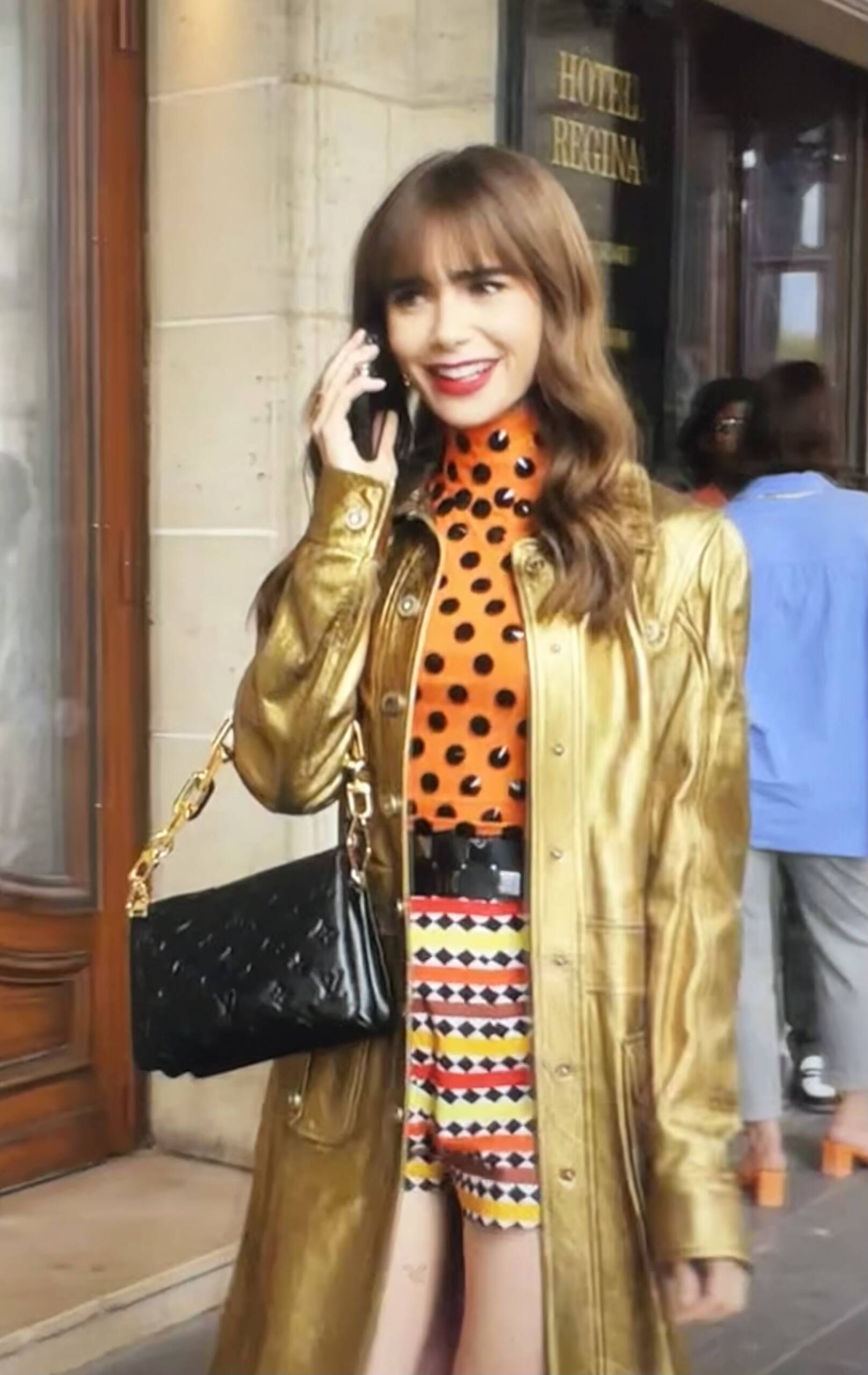 Emily in Paris Season 3 Episode 5 wearing a metallic gold button down overcoat, printed shorts, and orange and white polka dot turtleneck with a black Louis Vuitton BB Coussin bag. Lily Collins has her hair down and signature Birkin bangs.