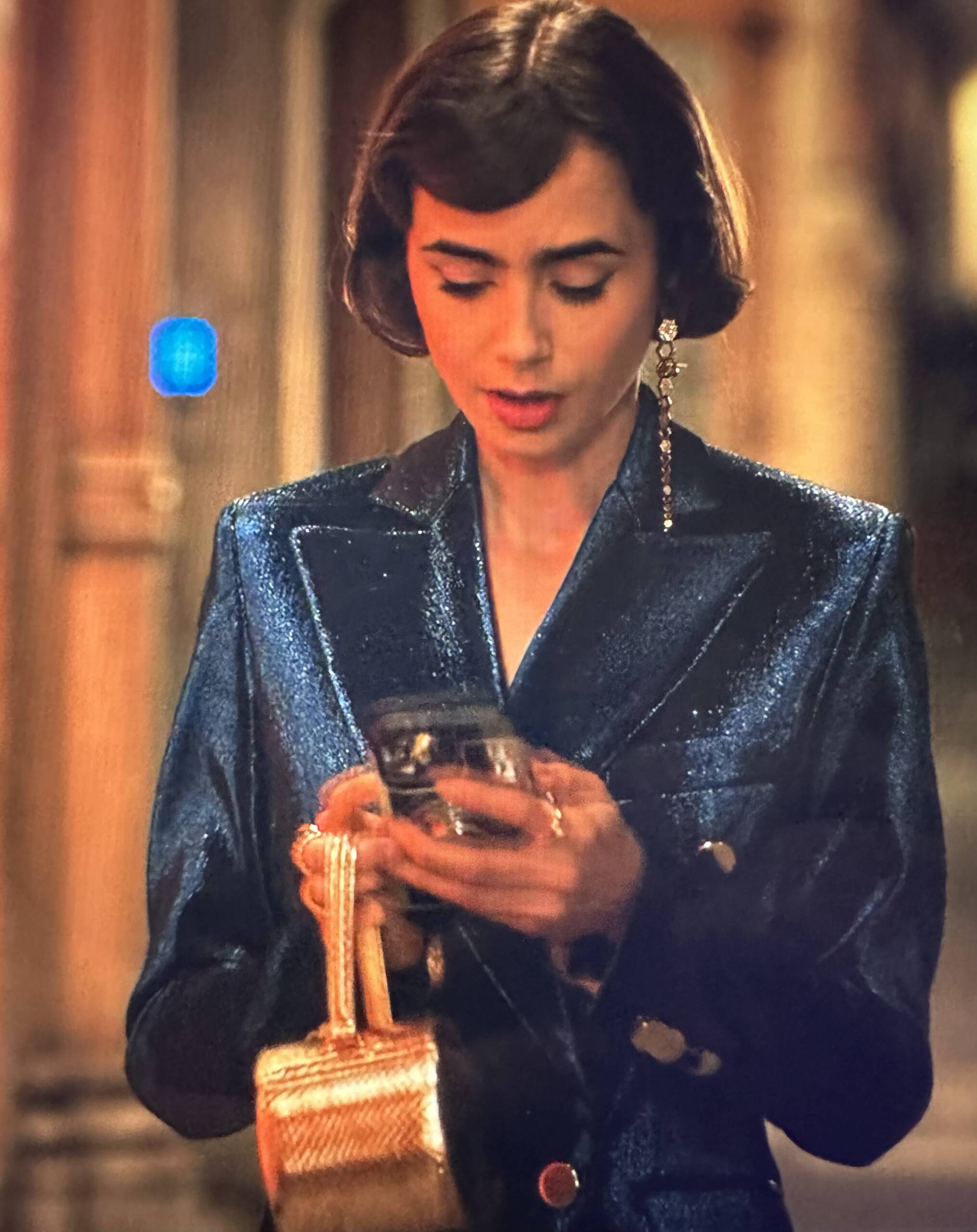 Emily in Paris Season 3 Episode 8, Lily Collins is wearing metallic blue jacket with short hair. She is carrying her signature camera phone case with a metallic gold round box triomphe python Celine mini bag. | Shop the fashion and handbags of Emily in Paris Season 3