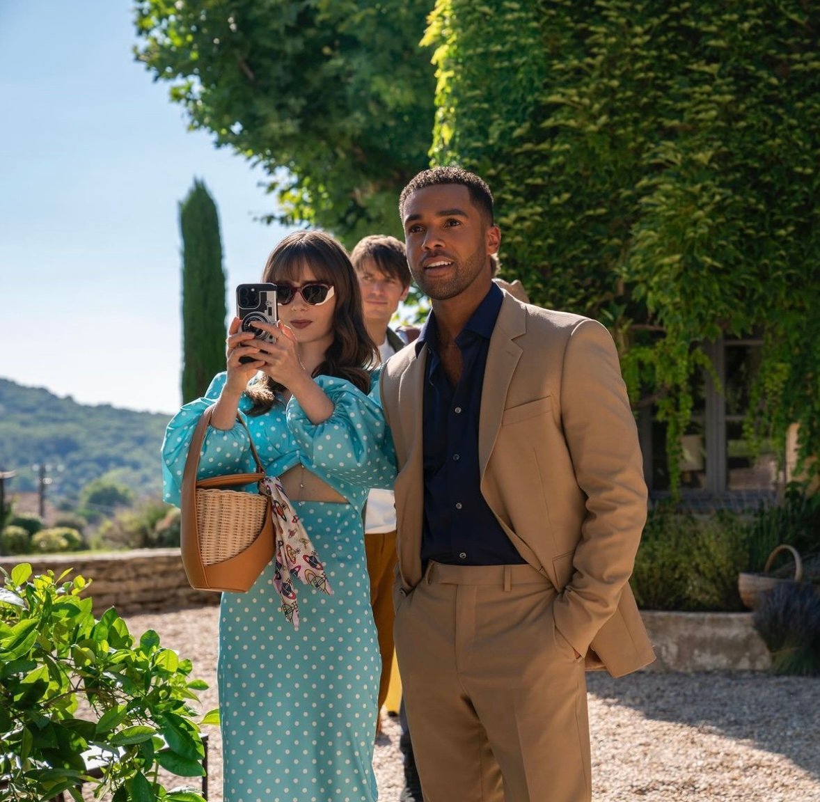 Emily in Paris Season 3 Episode 6, Lily Collins is wearing a blue polka dot long sleeve dress at the lavender farms in Provence, France. She is carrying a Polene Yke Bag in a tan color with a scarf tied to the side of the handle.