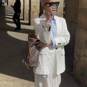 Blogger wearing a white suit with blue shirt and a Louis Vuitton Monogram Neverfull bag on one shoulder.