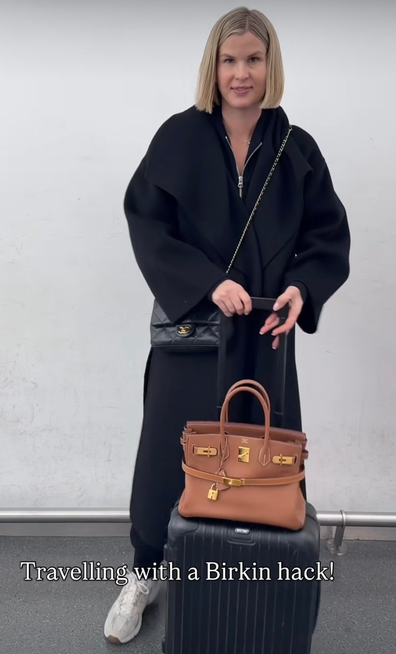 a woman showing the kelly belt travel hack, using a hermes kelly belt in gold to secure a gold birkin to her luggage.