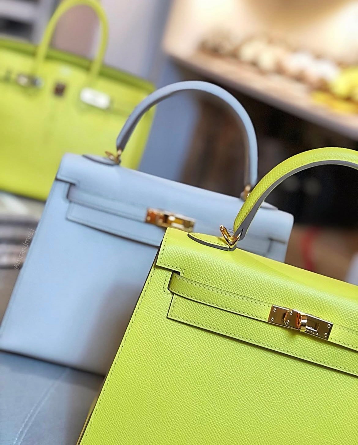 Close-up of two Hermès Kelly handbags—one in bright lime green and the other in pale blue—showcasing their gold hardware and elegant stitching. A third lime green Birkin bag is slightly blurred in the background, displayed in a luxury boutique setting.