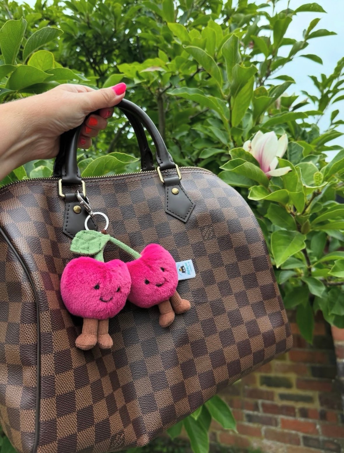 Hand holding a Louis Vuitton Damier Ebene Speedy bag with dark brown checkered pattern, featuring a plush cherry keychain, against a background of green foliage and a blooming magnolia flower.