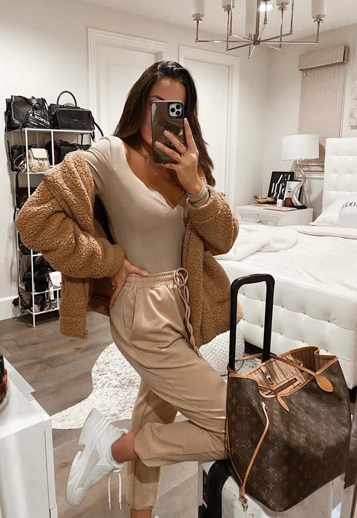 A person taking a mirror selfie in a softly lit bedroom with a Louis Vuitton Neverfull bag on top of a suitcase. She's dressed in a cozy neutral-tone outfit: a beige ribbed bodysuit tucked into matching drawstring joggers, layered with a plush, caramel-colored teddy jacket. On their feet are chunky white sneakers. They hold their phone up to obscure part of their face. In front of them sits a rolling suitcase topped with a large monogrammed designer tote bag. The bedroom features a tufted white bed, nightstand with chic décor, and a neatly organized bag display in the background.